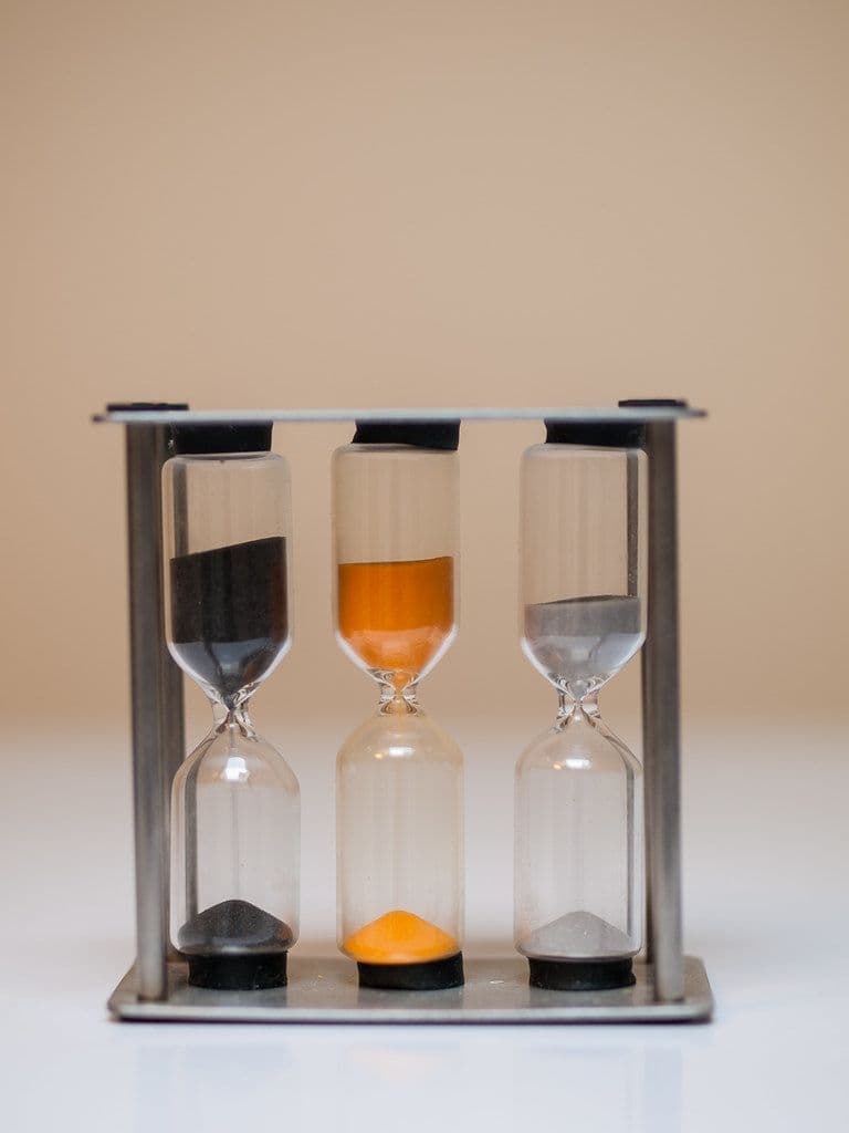 Three sand timers holding black, orange and grey sand standing side by side in a metal frame
