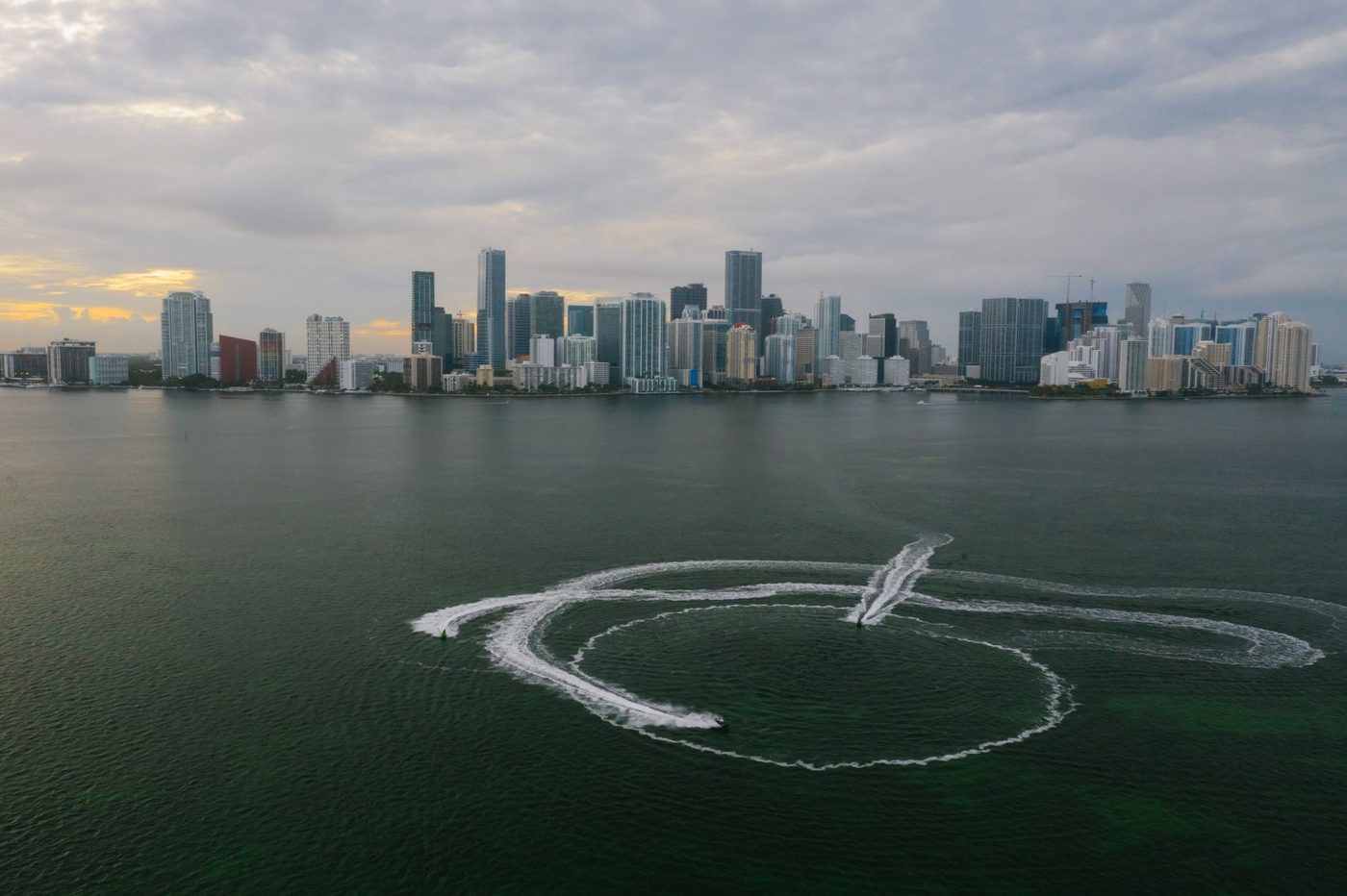The downtown Miami skyline seen across Biscayne Bay under heavy cloud, with two jet skis carving circles in the water