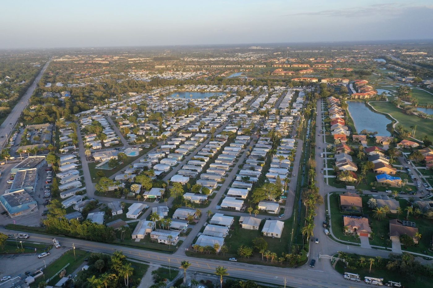 Aerial view at golden hour of a Florida community of low white single-storey homes laid out in rows, bordered by a golf course and retention ponds