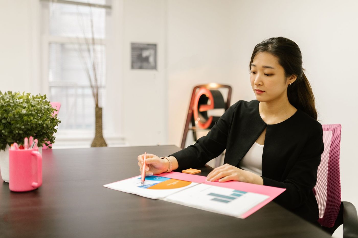 A woman in a black jacket sits at a dark desk reviewing a printed report with charts, a pen in her hand