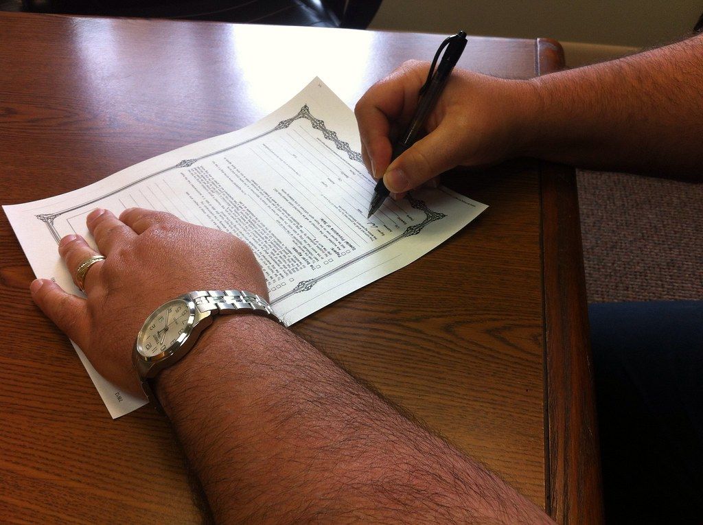 A man's hands, one wearing a metal watch, signing a printed form with a pen on a wooden table