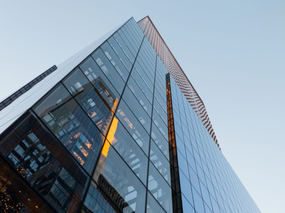 Looking up at the corner of a glass office tower against a pale sky, its windows reflecting neighbouring buildings at dusk