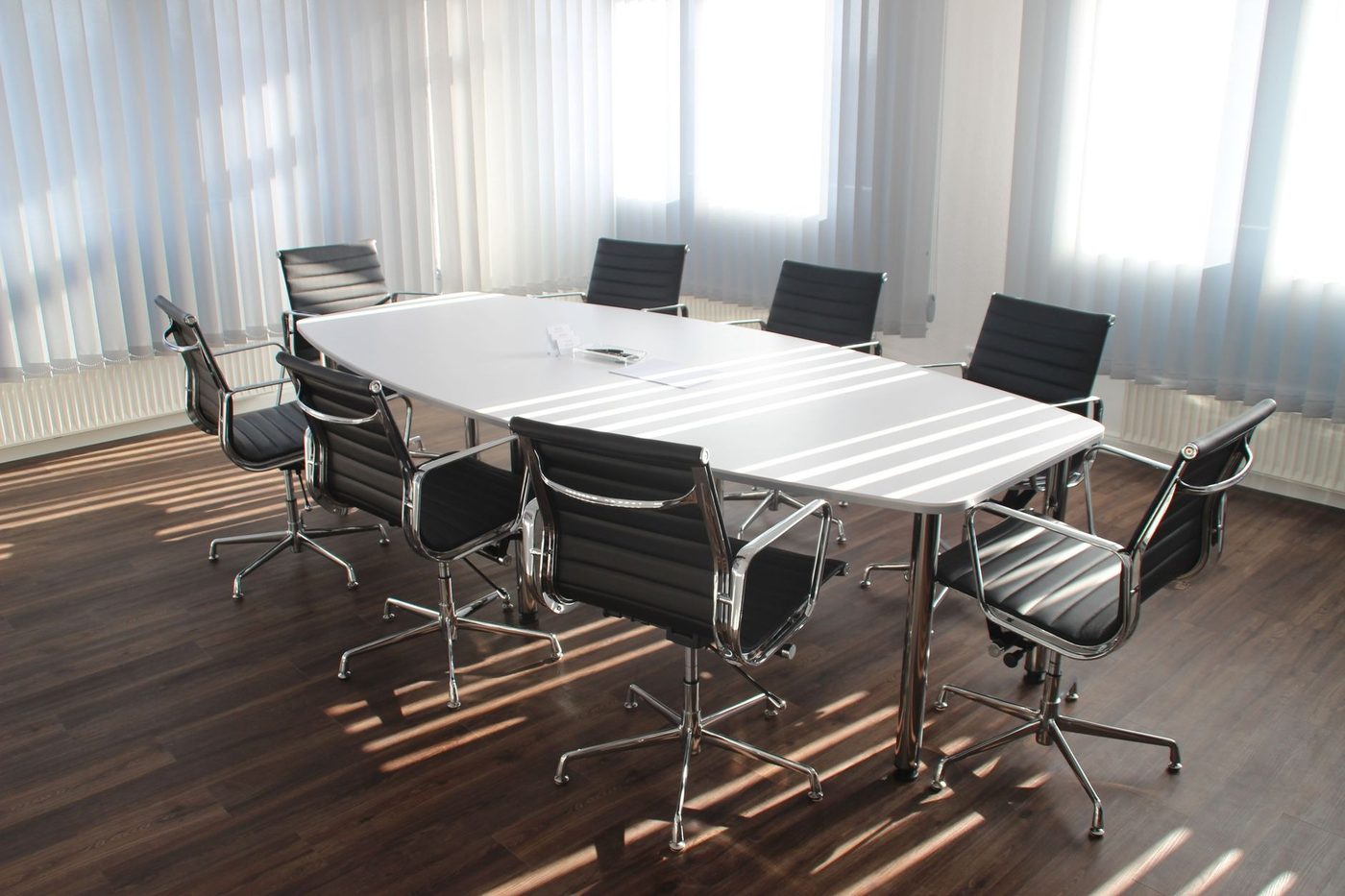 An empty boardroom with a white oval table and eight black leather chairs, sunlight falling across a dark wood floor through vertical blinds