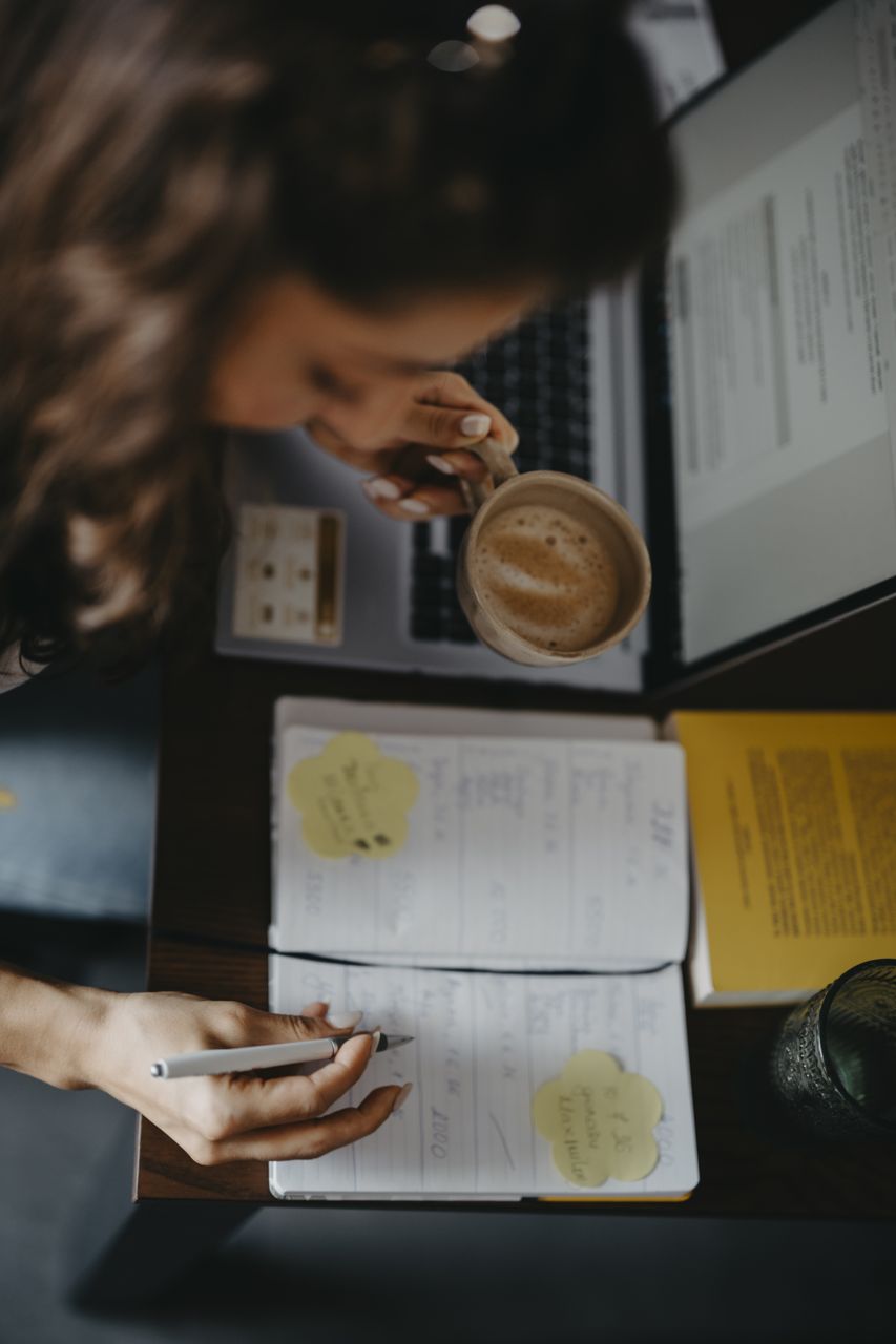 Overhead view of a woman holding a cup of coffee while writing figures in a ruled notebook beside an open laptop, yellow sticky notes on the page