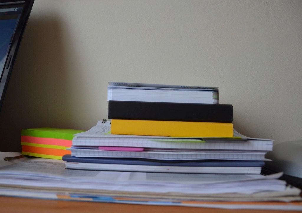 A stack of notebooks, folders and loose printed sheets piled on a desk beside a laptop and a block of coloured sticky notes