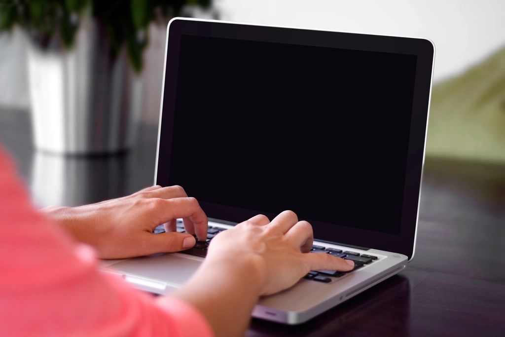 A person in a coral top typing on a silver laptop with a blank screen, a potted plant blurred in the background