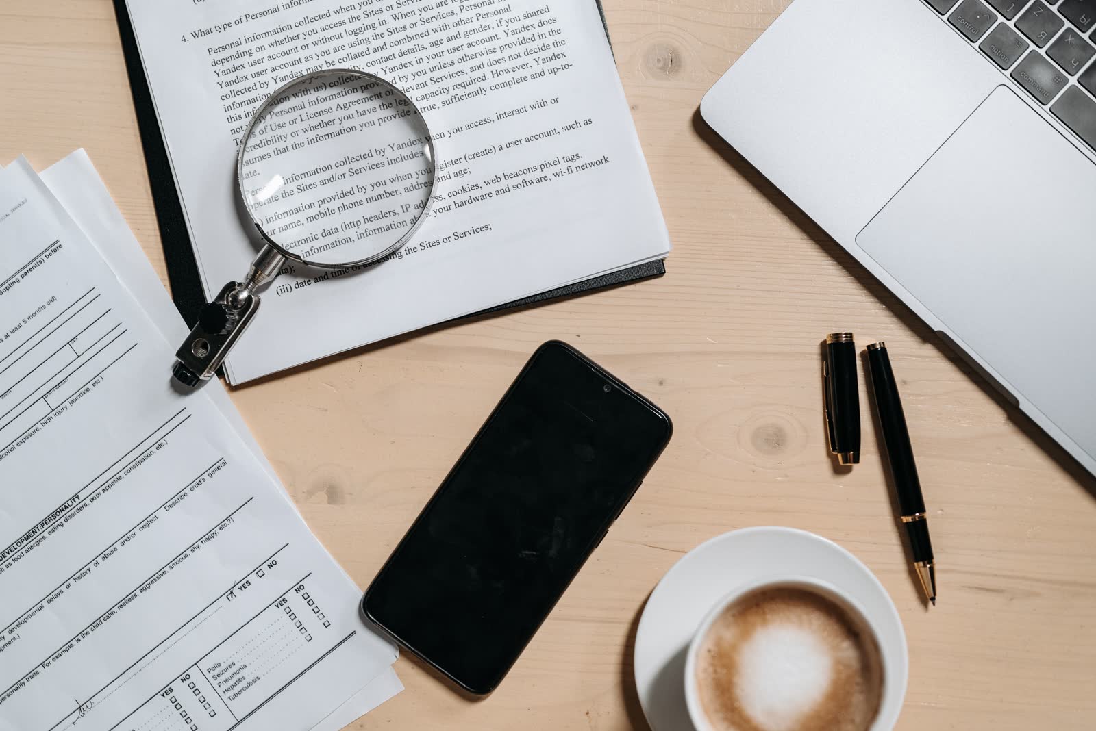 Printed documents, a laptop and a magnifying glass on a desk, seen from above