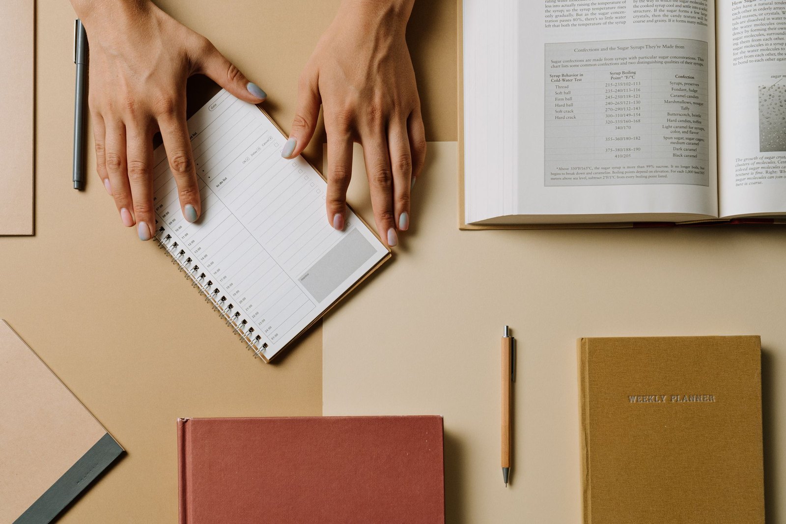 Hands writing in a small notebook beside an open book and bound journals
