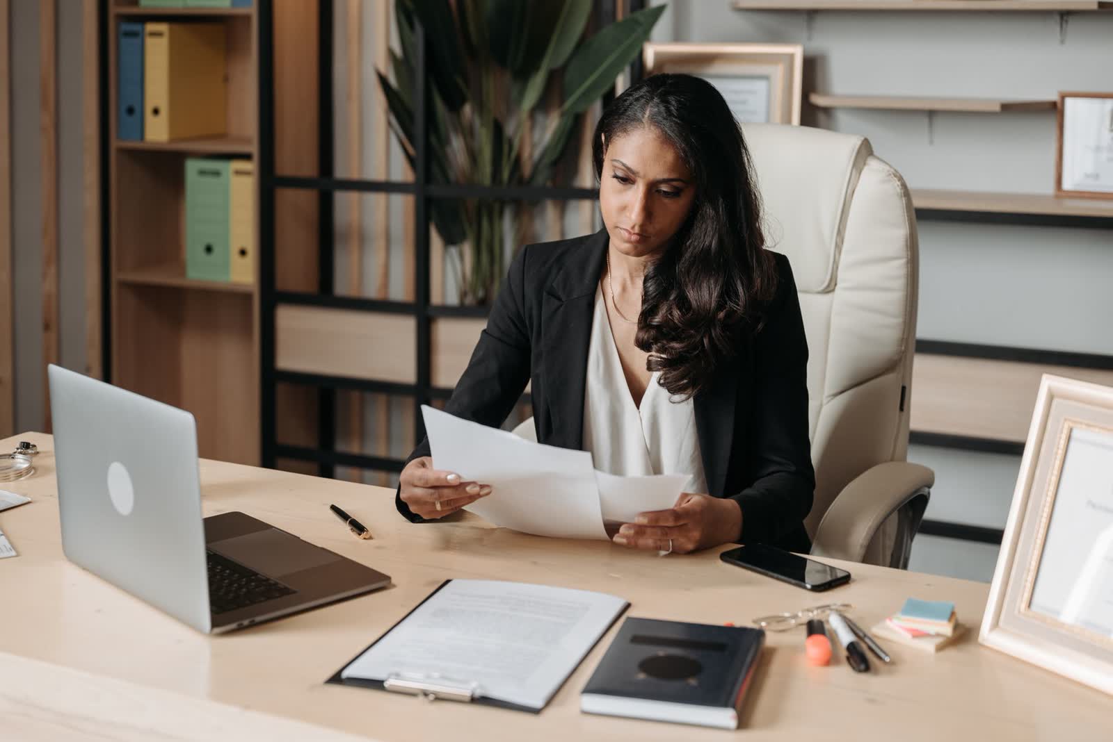 Businesswoman at an office desk reading a printed document beside a laptop
