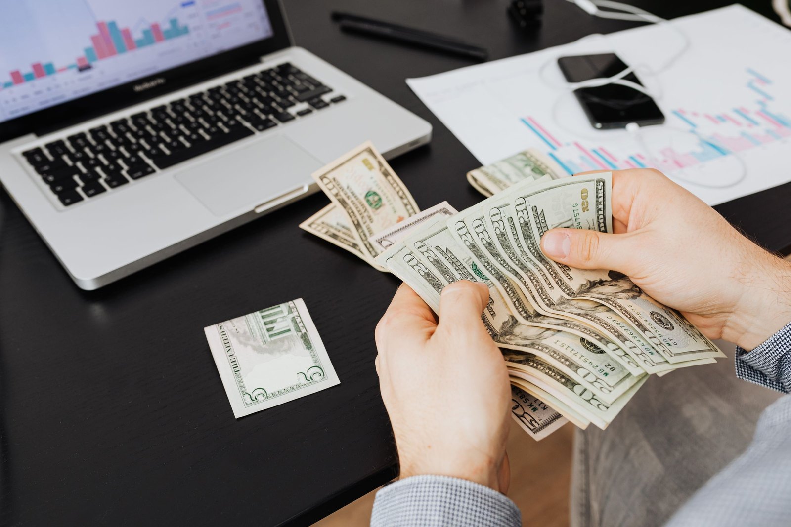 Person counting United States banknotes beside a laptop and a printed chart