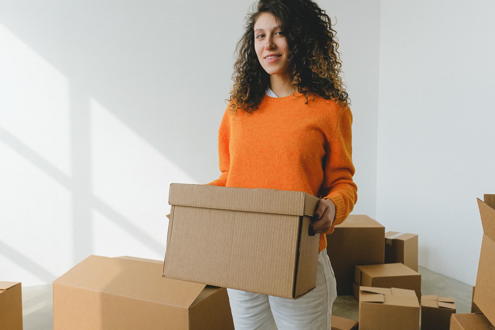 Woman carrying a cardboard box through a sunlit room stacked with boxes